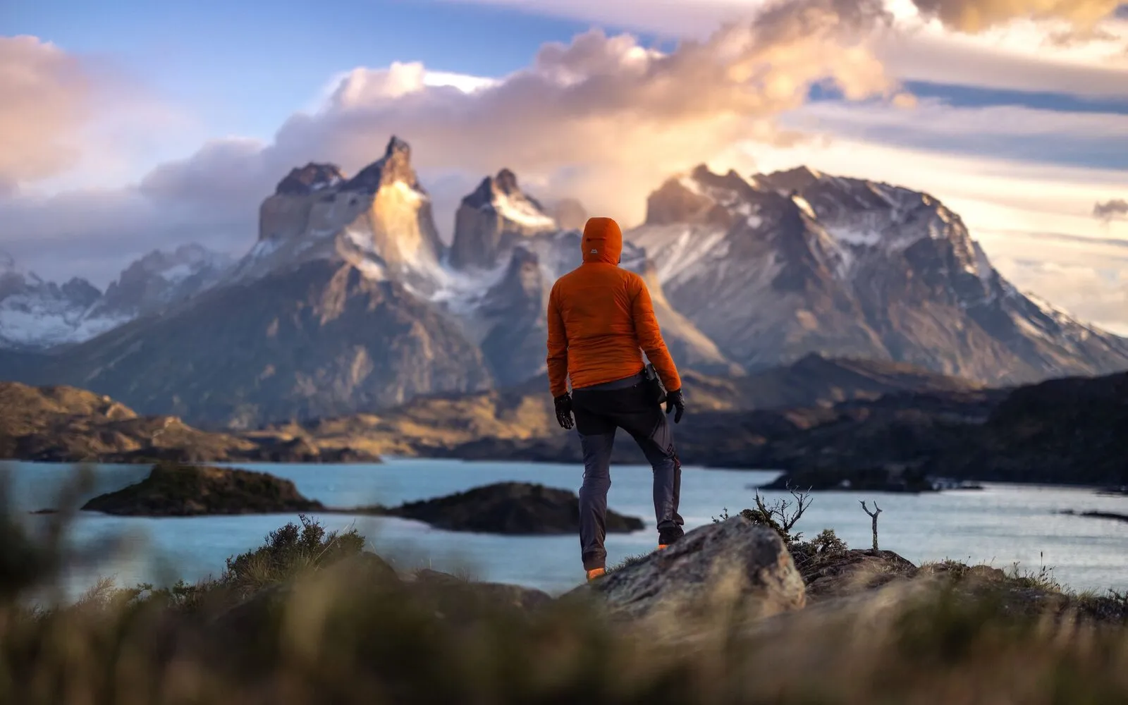 A hiker in an orange jacket looks out at the Torres del Paine peaks over a turquoise lake at sunrise